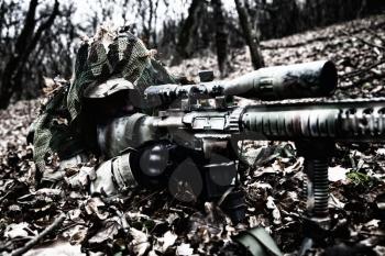 Army elite forces sniper, tactical group marksman, airsoft player lying on ground in forest, hiding in autumn foliage, covering himself with camouflage cape, searching targets through optical sight