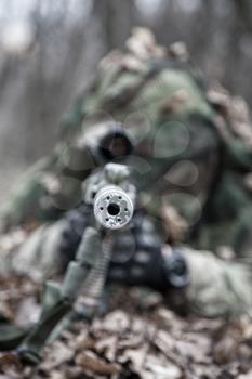Close up image of sniper rifle silencer muzzle, front view. Army elite forces shooter lying on ground in forest. Commando sniper hiding, masked himself in dry leaves. Marksman shooting from ambush