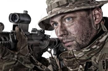Close-up studio portrait of army elite forces soldier, commando fighter, special operations squad shooter in camo bonnie, face paint camouflage, aiming service rifle, observing area with optical sight