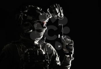Side view portrait of army soldier, modern combatant, special forces fighter in helmet, night-vision device, radio headset, hiding identity behind mask, armed service pistol, low key studio shoot