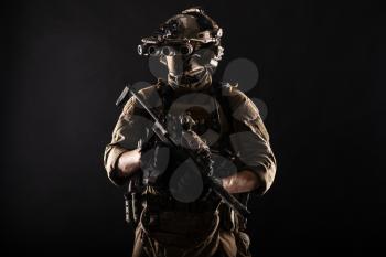 Army elite soldier with hidden behind mask and glasses face, in full tactical ammunition, equipped night vision device, radio headset, looking aside, armed short barrel rifle studio contour shot