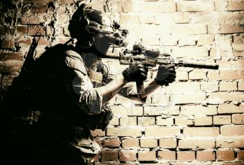Anti-terrorist squad fighter, army elite forces soldier in combat uniform and tactical ammunition, armed mini submachine gun, wearing night-vision device, moving along brick wall during sqb operation