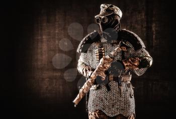 Post apocalyptic soldier in black mask and glasses, wool field cap and handmade armor from car tires and hauberk, standing at attention with submachine gun on shoulder, black background studio shoot