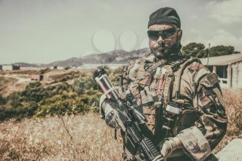 Navy SEALs fighter in ballistic goggles, equipped military ammunition and body armour, holding service rifle, looking in camera while standing outdoors. Special forces soldier half-length portrait