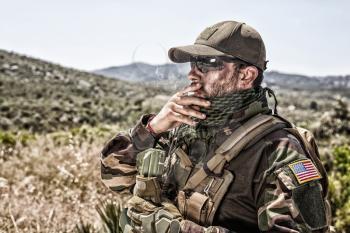 Army soldier, commando shooter in ammunition and battle uniform, standing in mountain area, looking into distance and smoking cigarette. Special operations forces fighter during mission