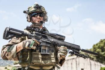 Half-length portrait of special forces sniper or marksman, army soldier in camouflage uniform and helmet with radio headset, standing on background of blue sky, holding sniper rifle with optical scope