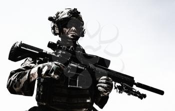 Half-length portrait of special forces sniper or marksman, army soldier in camouflage uniform and helmet with radio headset, standing on background of blue sky, holding sniper rifle with optical scope