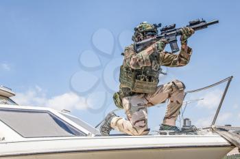 Army special forces soldier, commando fighter in full ammunition, wearing body armor and helmet, armed service rifle, standing on bow of speed boat, looking into distance during seacoast patrolling