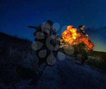 Army soldiers loaded with ammunition, running on battlefield at night. Commando team rushing on road, attacking enemy under artillery fire. Combatants in action, powerful explosion on starry horizon