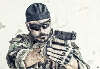 Army soldier, special forces fighter in camouflage uniform, ballistic glasses and cap, standing on background of white chipped wall, aiming and shooting with service pistol or handgun during fight