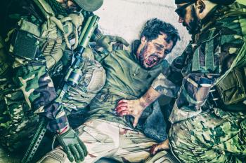 Commando fighters, Navy SEALs infantrymen giving emergency help to wounded comrade on battlefield. Soldier screaming in pain while teammate trying to stop bleeding from gunshot wound in his stomach