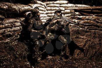 Soldiers covering from enemy fire in trench. Two Navy SEALs fighters, infantrymen in camouflage uniform and helmet, armed service rifles, hiding behind sandbags during night attack or gunfight