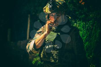 Commando soldier in camouflage uniform, ballistic glasses and bonnie, inhale cigarette smoke, smoking tobacco or marijuana joint in forest or jungles at night. Army infantryman relaxing after fight