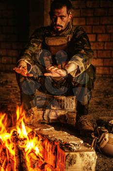 Special forces soldier after the fight sitting by the fire in ruined building warming his hands
