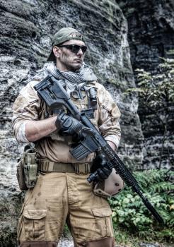 Private military contractor PMC in baseball cap with assault rifle in the rocks