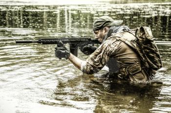Private military contractor PMC in baseball cap during river raid in the jungle waist deep in the water and mud