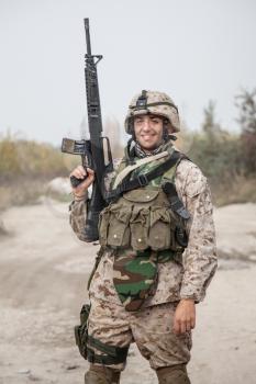Smiling army soldier, United States Marine Corps infantry shooter in camo battle uniform, protected with body armor and helmet, posing with assault rifle in hands while standing near country road