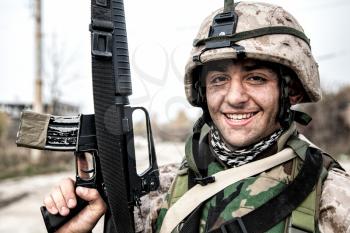 Smiling army soldier, United States Marine Corps infantry shooter in camo battle uniform, protected with body armor and helmet, posing with assault rifle in hands while standing near country road