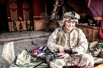 Marine Corps machine gunner smiling, disassembling, making maintenance of service weapon, checking gun barrel after cleaning at improvised outpost in abandoned building