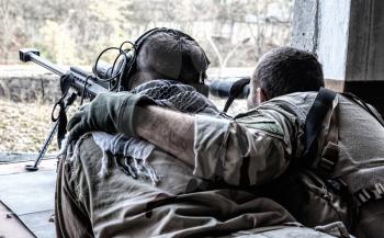 Military snipers team spotter observing battlefield, searching targets with binoculars, holding arm on shooters shoulder, correcting snipers fire with anti-materiel sniper rifle from hidden position