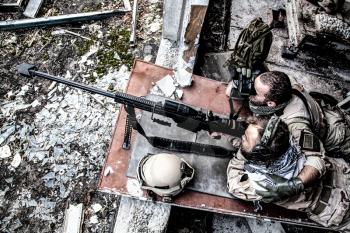 United States Navy SEAL sniper team firing with large caliber, anti material sniper rifle with optical sight from ruined, abandoned building. Elite members of anti-terrorist squad in modern warfare