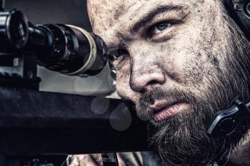 Army sniper with bearded, dirty face aiming with optical telescopic sight on sniper rifle, observing battlefield from ambush and searching targets to shoot, shooting on long range distances, close up