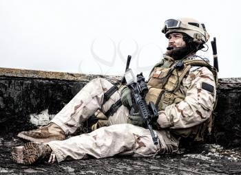 U.S. Navy SEAL infantryman, commando marksman in battle uniform, armed with assault service rifle with optics sight, observing territory while sitting on observation post on roof of ruined building