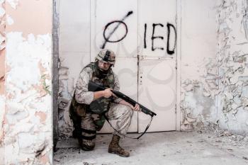 Army soldier, marine infantry, commando in camo uniform and helmet, armed with shotgun, standing on knee, hiding behind concrete column in ruined, abandoned building, during military firefight in city