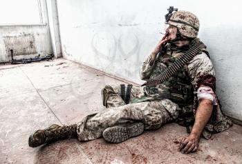 Soldier in combat uniform and ammo belt over chest, sitting on floor and smoking after being shoot in shoulder and put bandage on wounded hand, dropped weapon lying nearby with blood stains around