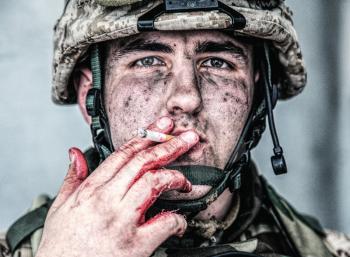 Close up portrait of wounded soldier, U.S. marine with dirty face, holding cigarette with soiled in blood fingers and smoking. Military medic resting after field operation. Traumatic war experiences