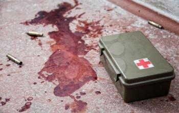 First aid kit metal box lying on floor with empty bullet shells and fresh stains of human blood around. Emergency medical care for gunshot wounds, bleeding stop, tactical combat casualty care concept