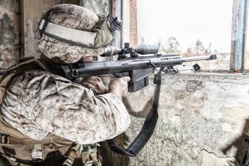 Marines sniper armed with large caliber, anti-materiel sniper rifle hiding in ruined urban building, shooting enemy targets on range from shelter, sitting in ambush. Military firefight in city