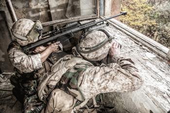 Marines sniper team armed with large caliber, anti-materiel sniper rifle hiding in ruined urban building, shooting enemy targets on range from shelter, sitting in ambush. Military firefight in city