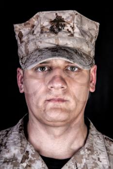 Close up studio portrait of U.S. marines infantry soldier in camouflage combat uniform and patrol cap with United States Marine Corps emblem on it, looking at camera, isolated on black background