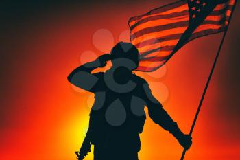 Silhouette of army soldier, armed service rifle, holding USA national flag, saluting on background of sunset or dawn sky. Military respect and honor, patriotism veterans and heroes remembrance