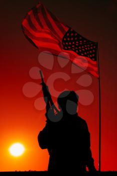 Silhouette of US army infantry soldier, special forces rifleman in combat helmet, armed assault rifle standing under waving United States of America national flag with setting sun on background