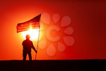 Silhouette of US army soldier, special operations shooter holding waving on flagpole national flag while standing on background of sunset. American army hero, United States or America military victory