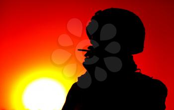 Silhouette of army soldier in combat helmet smoking cigarette on background of red sunset sky and setting sun backlight. Special operations forces fighter resting at end of day, relaxing after fight