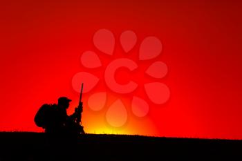 Silhouette of army sniper, special forces soldier, professional mercenary or hunter in bonnie hat, carrying backpack and sitting on top of hill with sniper rifle on background of sunset or dawn sky
