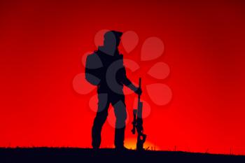 Silhouette of army special forces sniper standing with rifle on background of sunset sky. Commando fighter in boonie hat and ammunition patrolling area at nighttime. Hunter hunting on sunset or dawn