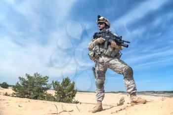 Portrait of United states airborne infantry corporal with arms, camo uniforms dress. Combat helmet on, tactical light, boots and kneepads, low angle view