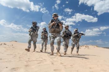 Team of United states airborne infantry men with weapons moving patrolling desert. Sand and blue sky on background of squad, sunlight, front view