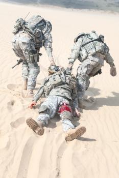 Team of airborne infantry paratroopers saving life of injured brother in arms dragging carrying him on desert sand. No man left behind