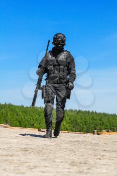 Swat police operator with sniper rifle in black uniforms walking towards camera. Blue sky and greenery background. Relaxing after mission