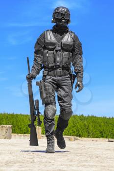 Swat police operator with sniper rifle in black uniforms walking towards camera. Blue sky and greenery background. Relaxing after mission