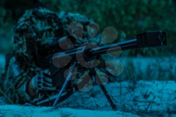 Army sniper with big rifle lying in wait in the forest at nighttime
