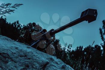 Army sniper with big rifle lying in wait in the forest at nighttime. Low angle view, diagonal shot
