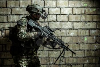 US Army Ranger member with machinegun and night vision goggles moving along the wall during mission