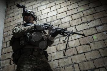 US Army Ranger with machinegun and night vision goggles moving along the wall during mission. Low angle view