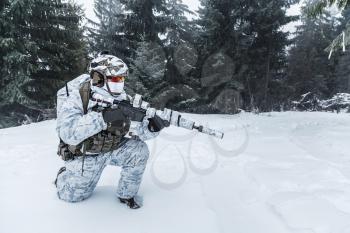 Winter arctic mountains warfare. Action in cold conditions. Trooper with weapons in forest somewhere above the Arctic Circle on one knee
